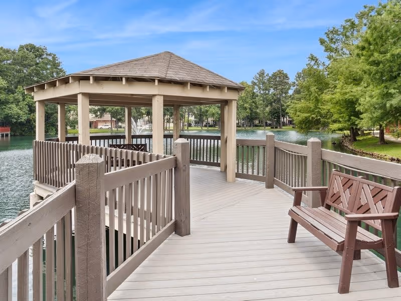 Wooden lakeside gazebo and boardwalk with a bench overlooking a pond and fountain surrounded by trees.