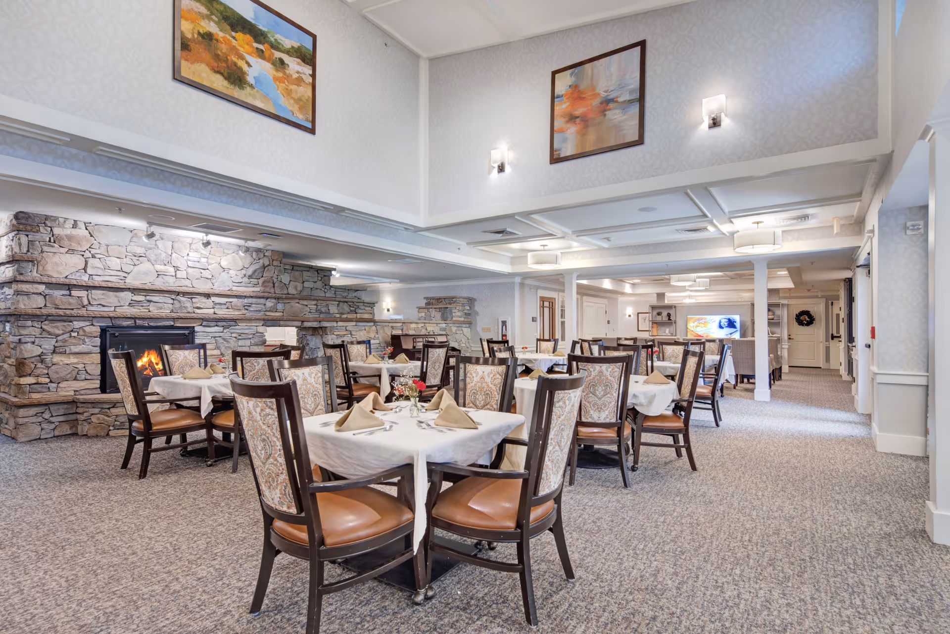 A spacious dining room with multiple tables covered in white tablecloths, each set with beige napkins and small floral centerpieces. The room features a stone fireplace with a fire burning, patterned upholstered chairs, carpeted floor, and soft lighting. Two framed paintings hang on the walls above the fireplace and seating area.