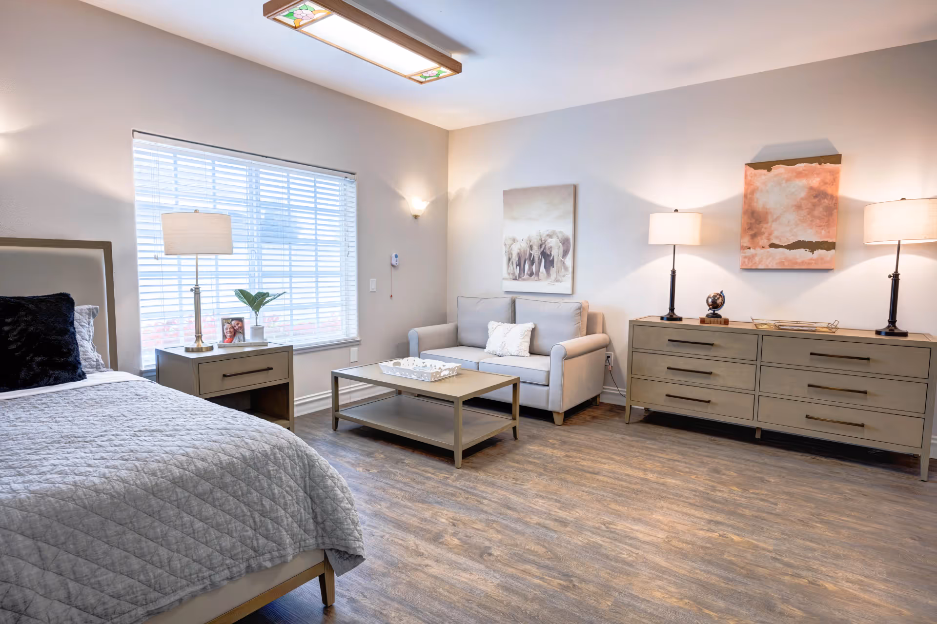A cozy bedroom with a bed covered in a gray quilt, a nightstand with a lamp and a small plant, a window with blinds, a small gray loveseat with a white pillow, a coffee table, and a dresser with two lamps and decorative items. The walls are light-colored with two pieces of artwork hanging above the loveseat and dresser.