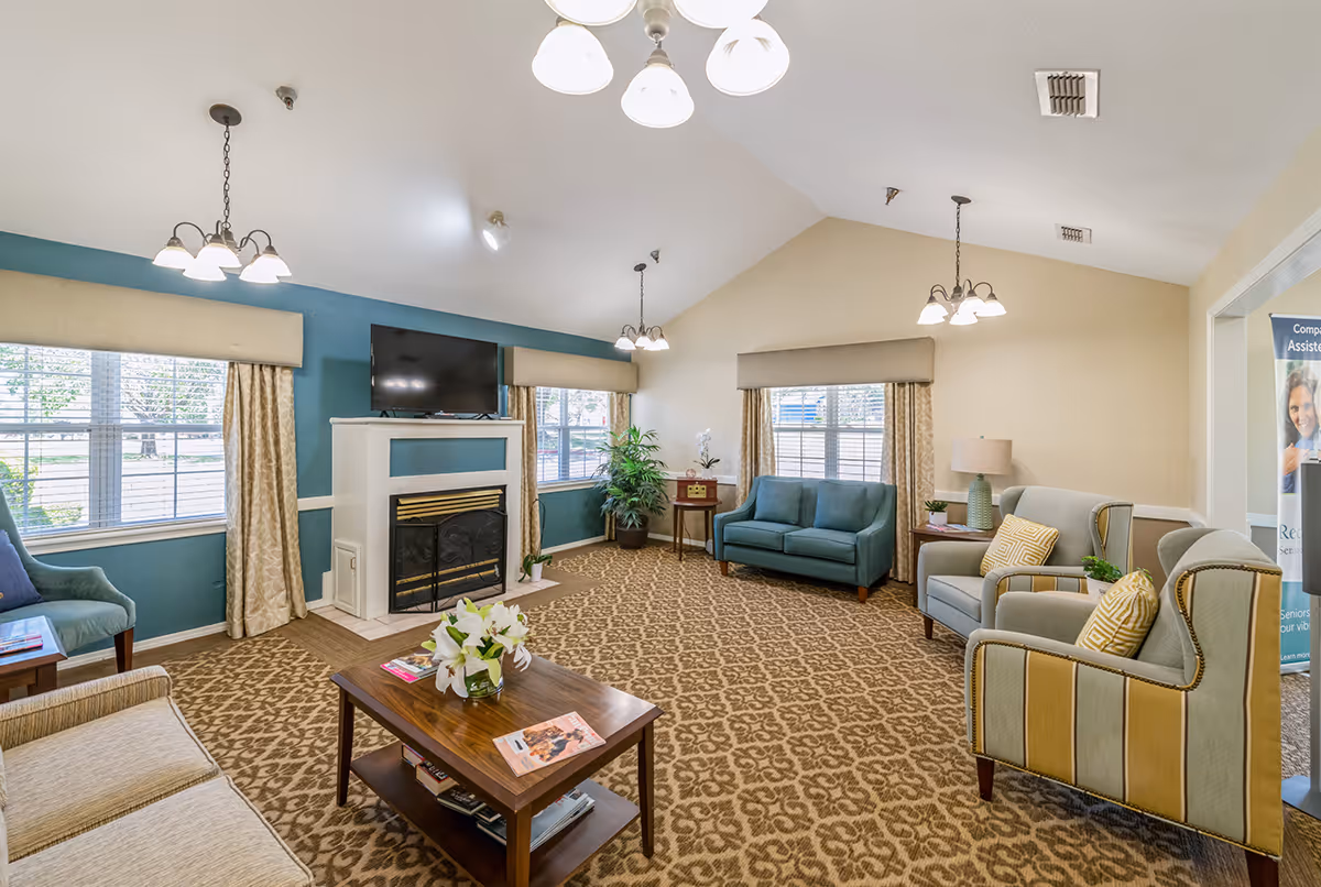 Well-lit communal living room with sofas, armchairs, a fireplace with TV above, coffee table, and patterned carpet.
