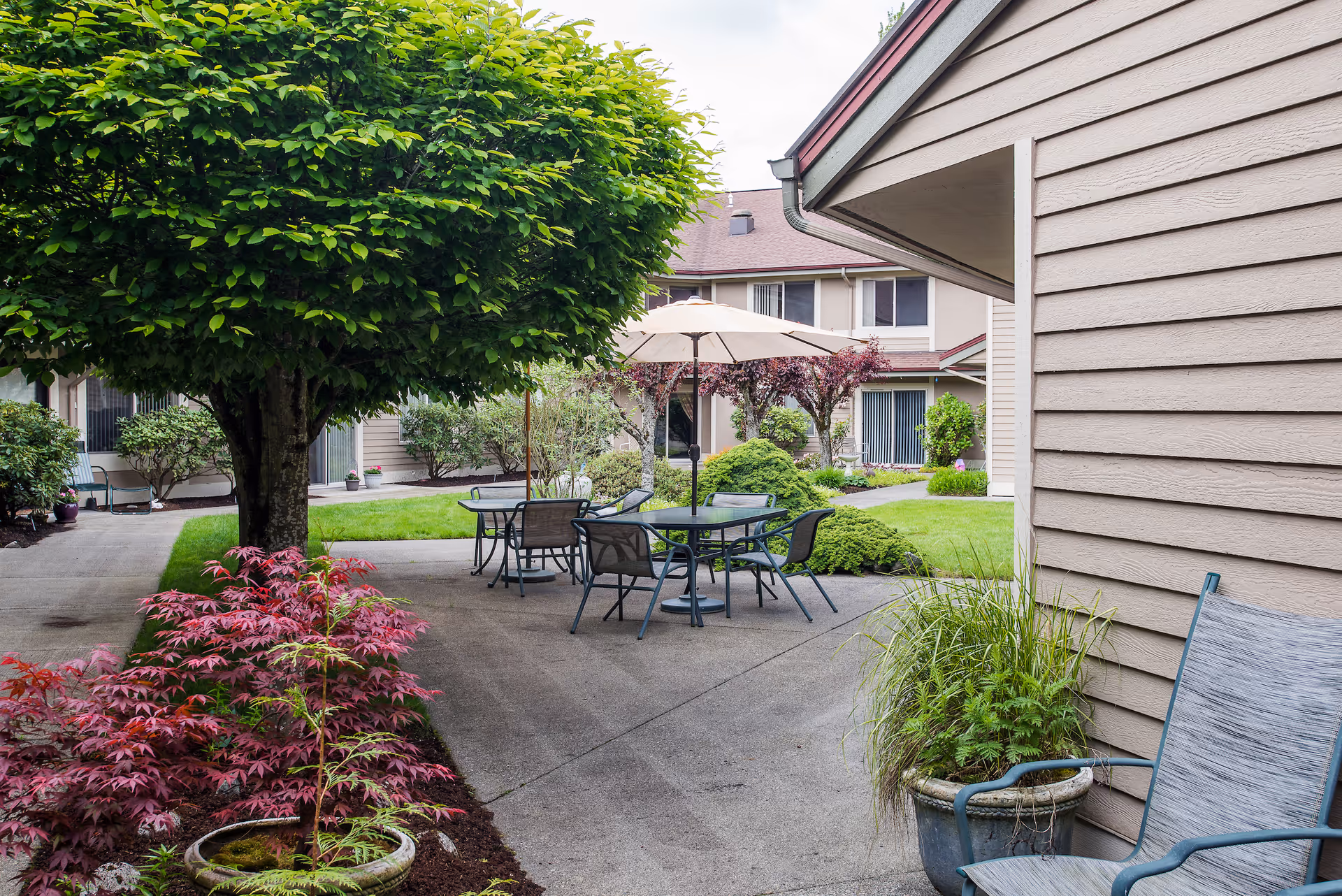 A landscaped courtyard with patio tables and chairs under an umbrella, surrounded by shrubs, trees, and residential building siding.