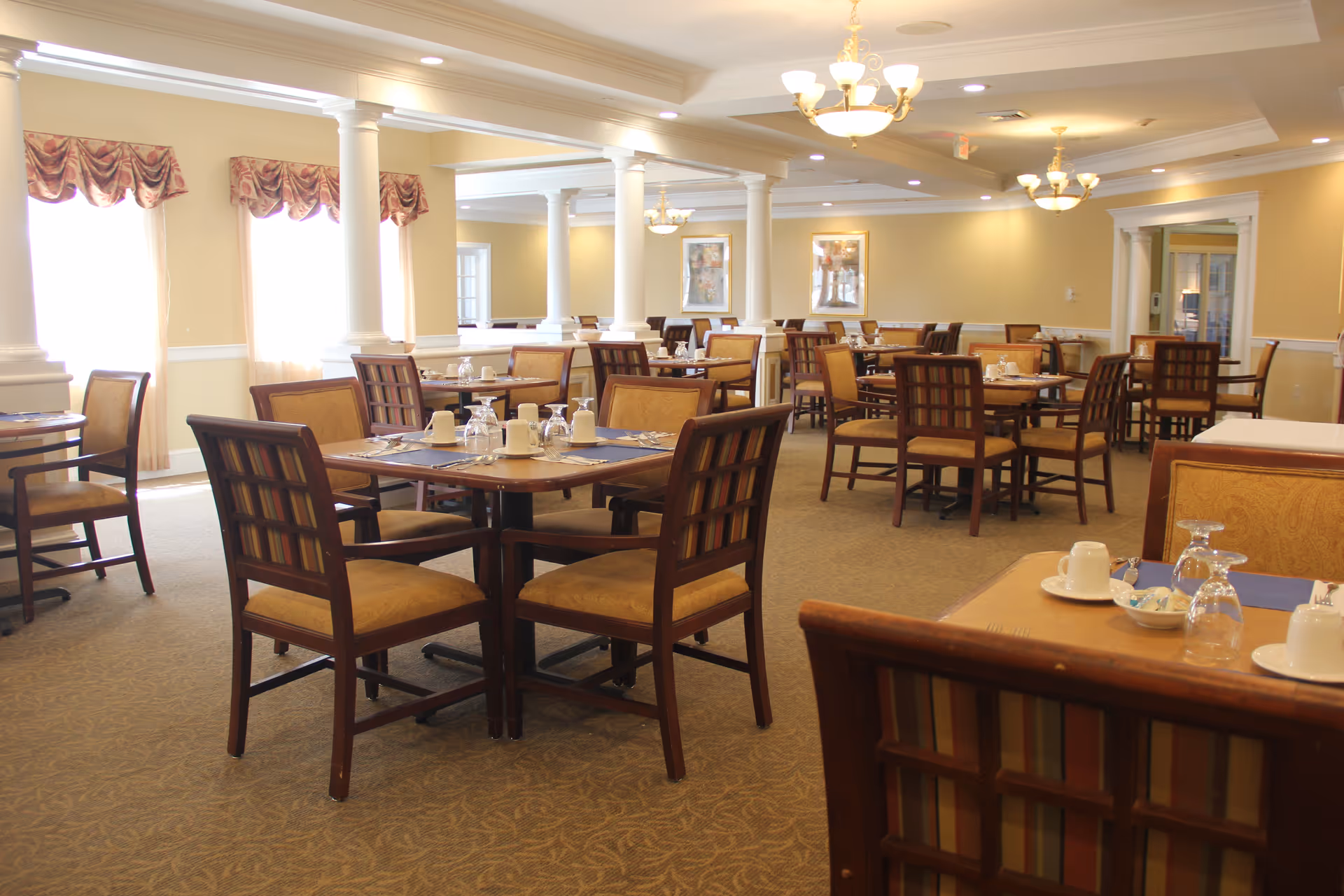 A spacious dining room in a senior living facility with multiple wooden tables and chairs arranged neatly. Each table is set with cups, glasses, and napkins. The room is well-lit with natural light coming through large windows with decorative valances, and ceiling chandeliers provide additional lighting. The walls are painted a soft yellow, and there are white columns and framed artwork on the walls.