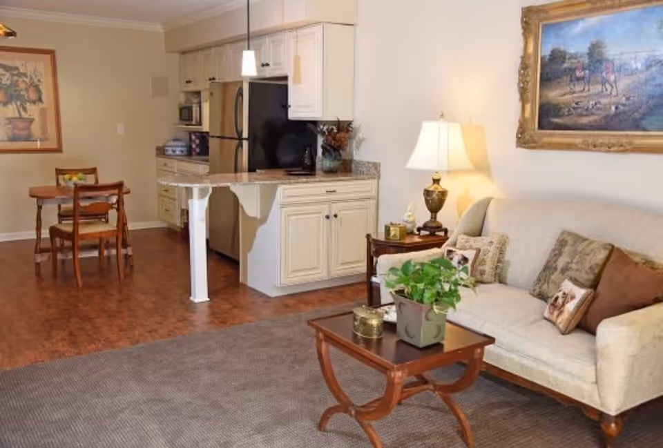 Interior view of a cozy assisted living apartment featuring a small living room with a beige sofa adorned with decorative pillows, a wooden coffee table with a potted plant, and a side table with a lamp. Adjacent to the living room is a compact kitchen area with cream-colored cabinets, a granite countertop, and stainless steel appliances. A small wooden dining table with two chairs is positioned near a wall with framed artwork.
