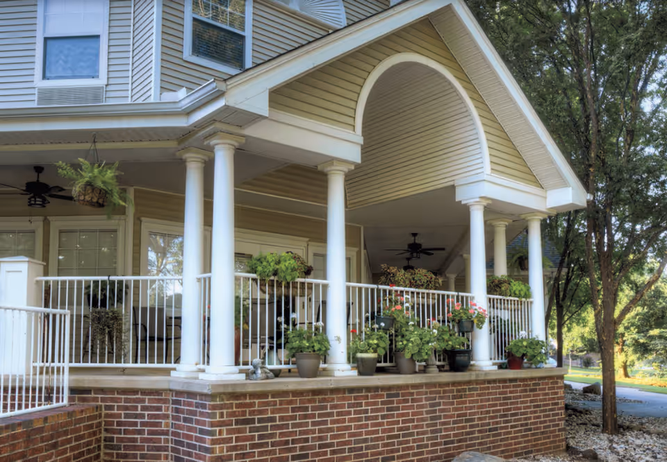 Covered front porch with white columns, railings, potted plants, and a brick foundation at the building entrance.