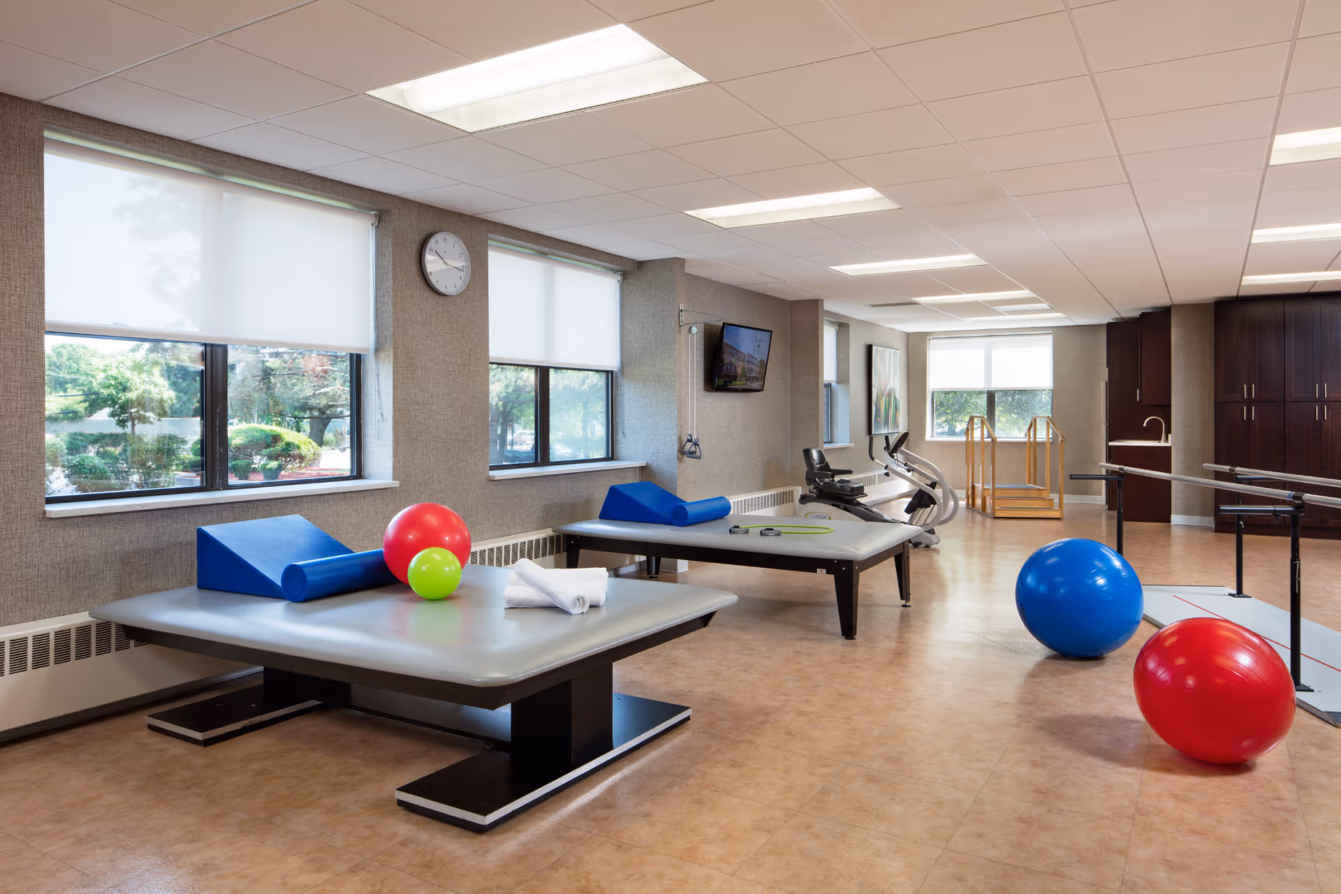 A spacious rehabilitation room with large windows letting in natural light. The room contains two padded therapy tables with foam rollers and exercise balls in red, blue, and green. There is a stationary exercise bike, parallel bars for walking exercises, and a wall-mounted TV. The floor is light brown, and the walls are neutral-colored with some artwork and cabinetry.