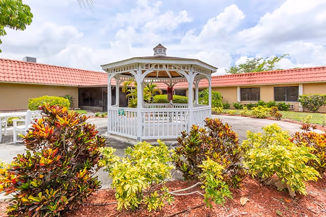 A white wooden gazebo surrounded by colorful bushes and plants in a courtyard area with single-story buildings with red-tiled roofs in the background under a partly cloudy sky.