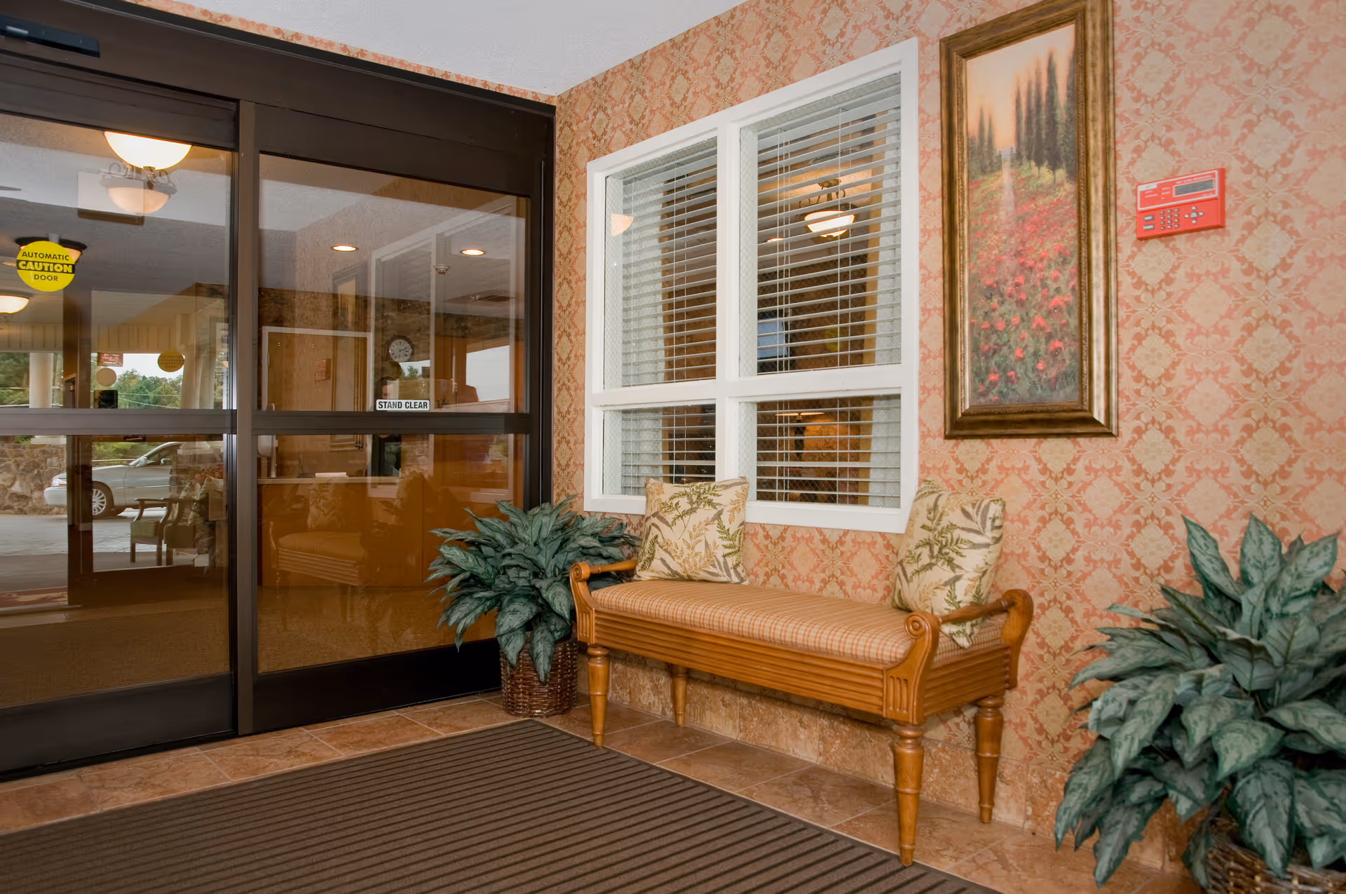Entrance area of a facility with automatic sliding glass doors, a wooden bench with two patterned cushions, two potted plants, a window with white blinds, a framed painting of a landscape on a patterned wallpapered wall, and a fire alarm panel.