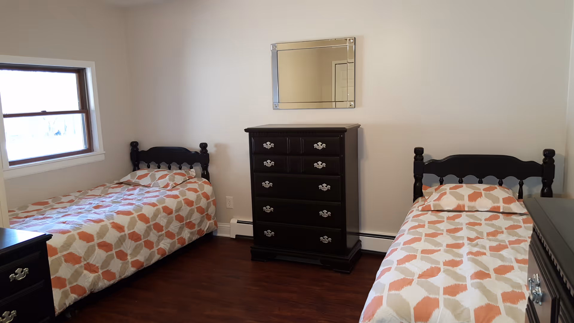 A simple bedroom with two twin beds placed against opposite walls, each with matching patterned bedspreads in shades of orange, beige, and white. Between the beds is a dark wooden chest of drawers with a rectangular mirror mounted on the wall above it. There is a window on the left wall letting in natural light, and the floor is a dark wood finish.