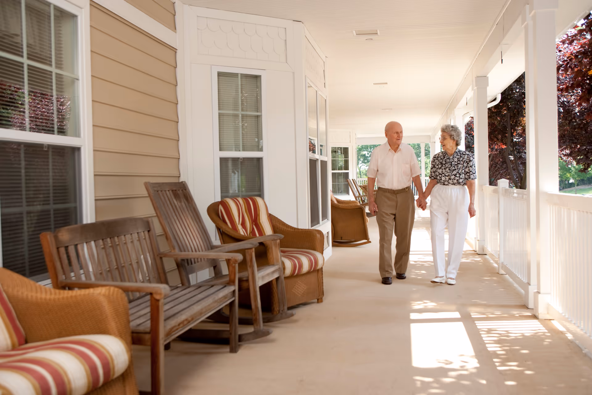 An elderly couple holding hands and walking along a covered outdoor porch with wooden and wicker chairs lined up against the wall. Trees and greenery are visible outside the porch.