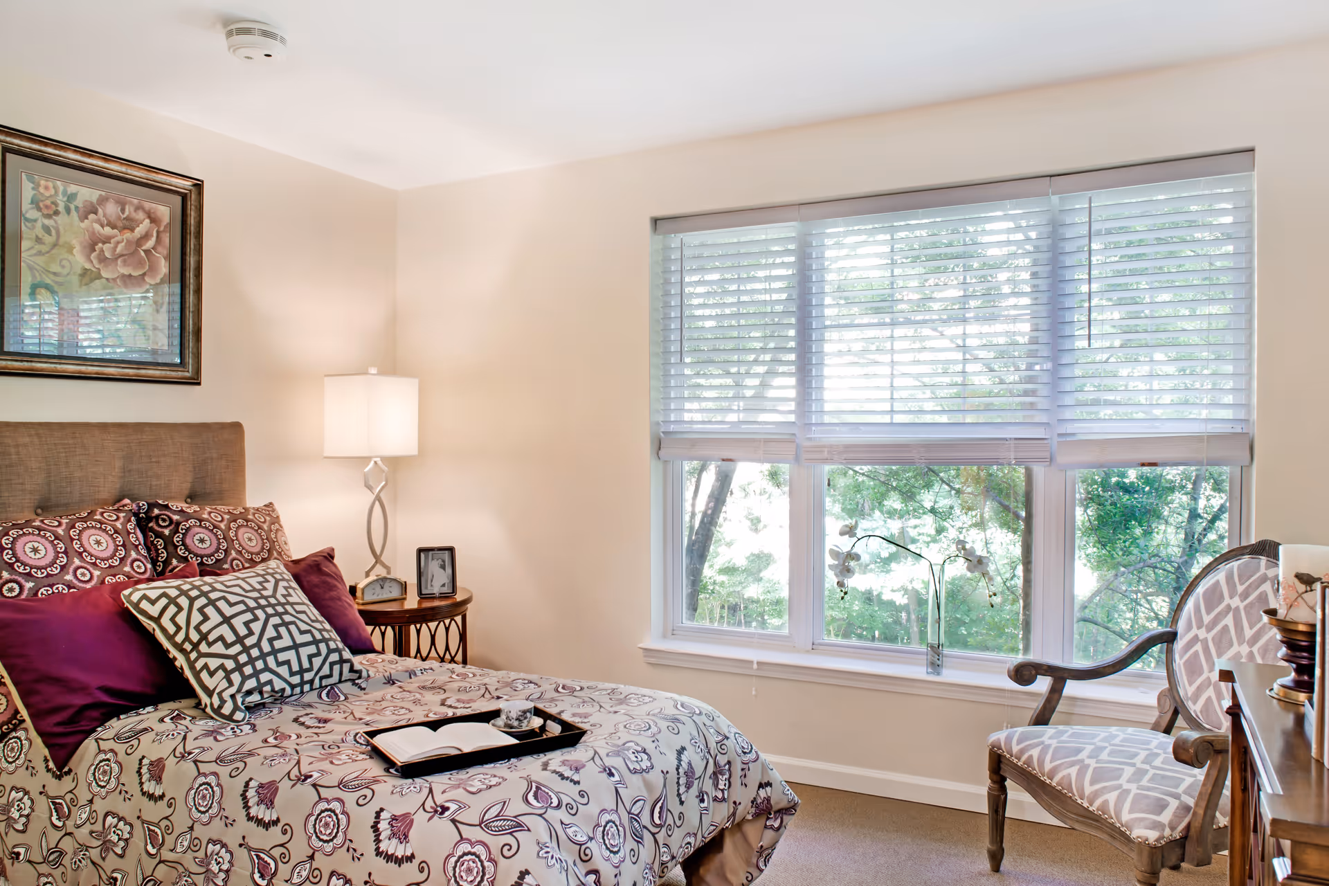 A cozy bedroom with a bed featuring patterned bedding and multiple pillows. A bedside table with a lamp, clock, and framed photo is next to the bed. Large windows with white blinds allow natural light to fill the room. A decorative chair with a patterned cushion is placed near the window, and a framed floral artwork hangs on the wall above the bed.