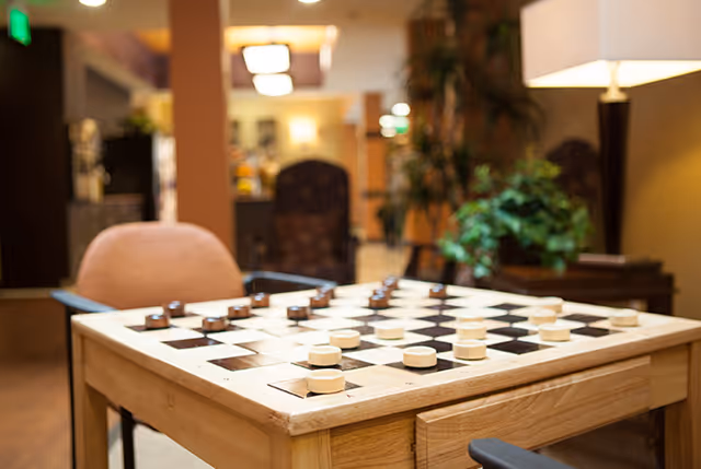 Wooden table with a checkers board and pieces in a cozy common area with chairs, a lamp, and plants in the background.