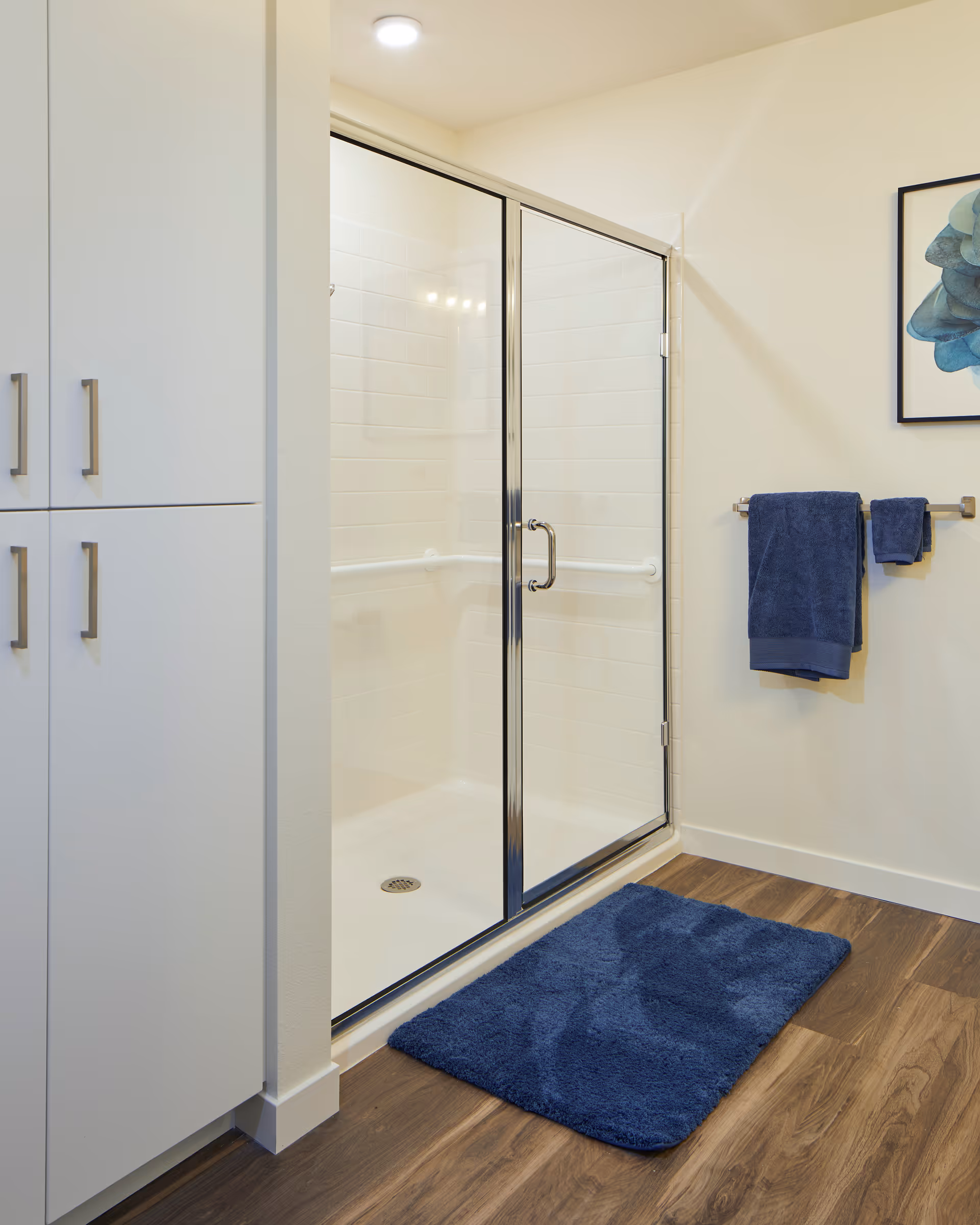 Glass-enclosed shower in a bathroom with white cabinetry, blue bath mat and towels, and wood-look flooring.