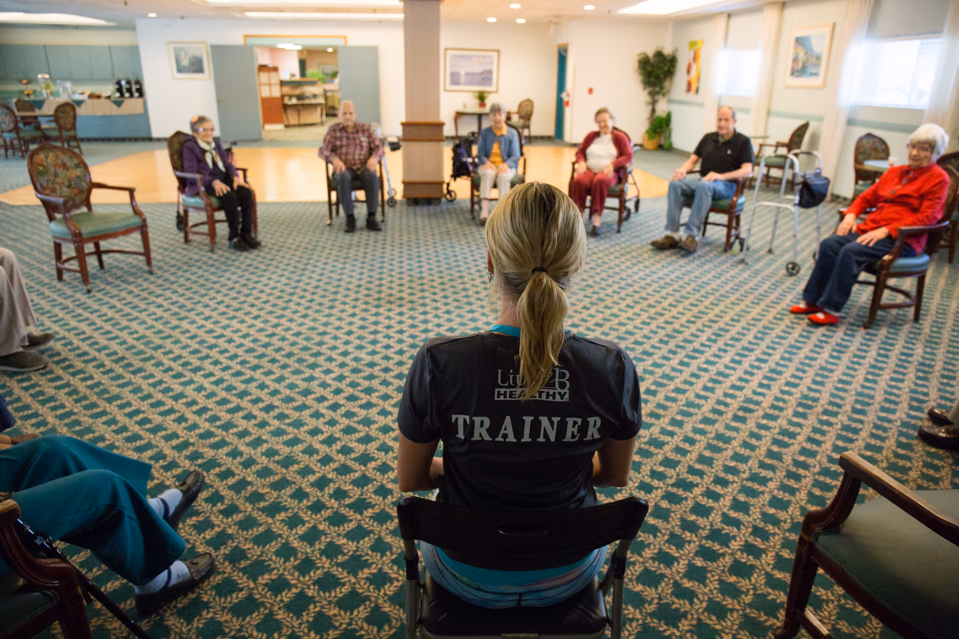 A trainer leads a seated exercise class with seniors arranged in a large carpeted common room.