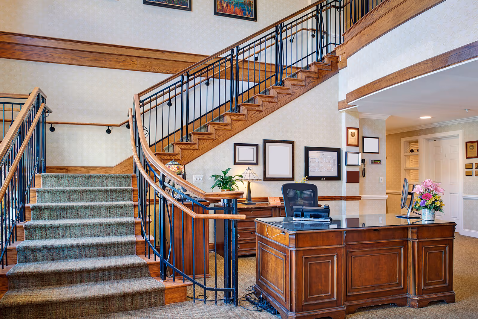 Interior view of a senior living facility lobby area featuring a wooden reception desk with a computer and a vase of flowers. Behind the desk is a staircase with wooden steps and black metal railings leading to an upper floor. The walls are decorated with framed pictures and certificates, and there is a small lamp and a plant on a side table.