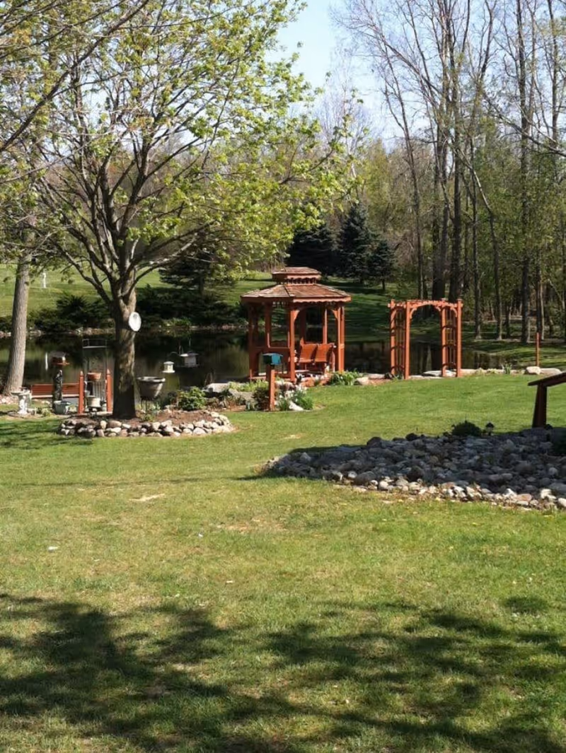 A peaceful outdoor garden area with green grass, a large tree with fresh leaves, a wooden gazebo, a wooden archway, and a small pond surrounded by trees and shrubs in the background.