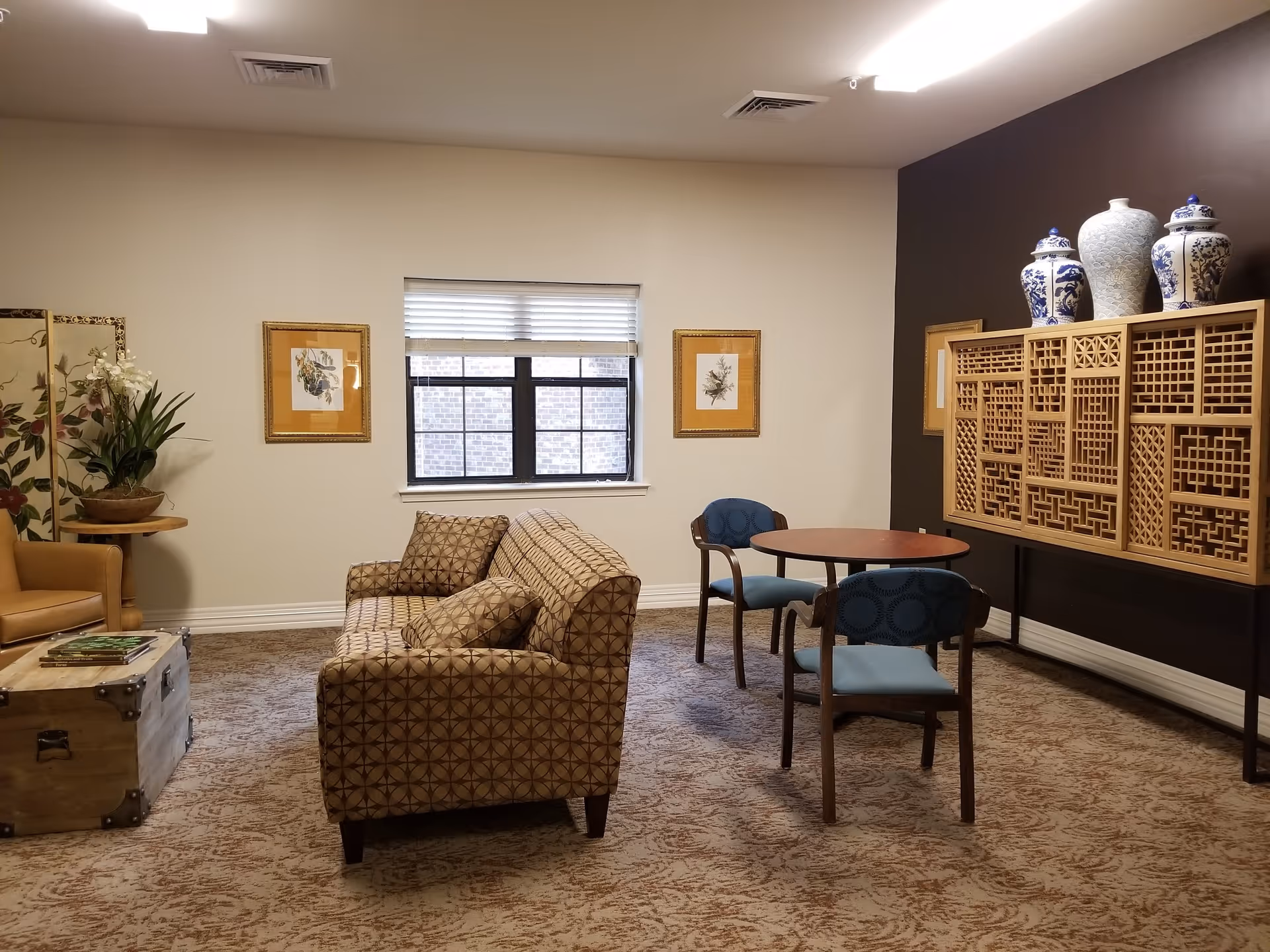 A cozy living room area with a patterned sofa and matching armchair, a wooden coffee table resembling a trunk, a small round table with two blue cushioned chairs, decorative vases on a wooden cabinet against a dark accent wall, two framed botanical prints on the wall, a window with blinds, and a potted plant on a small round side table.