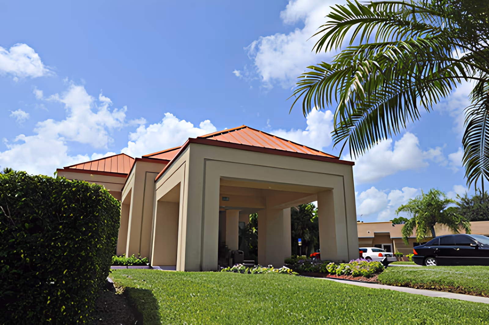 Entrance canopy of a beige building with a red roof, palm trees, parked cars, and landscaped lawn under a blue sky.