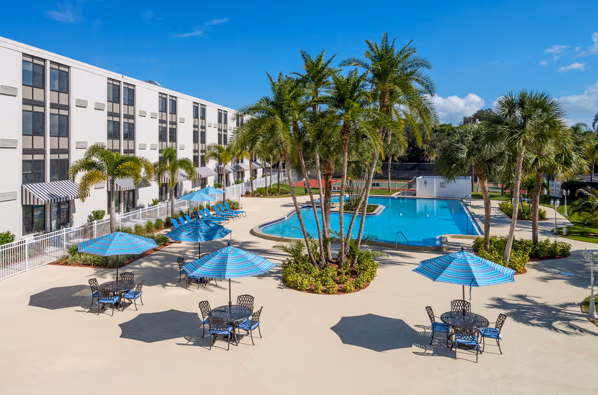 Outdoor pool area at a senior living facility with several blue and white striped umbrellas shading black metal tables and chairs. There are palm trees planted around the pool and along the building, which is a multi-story white structure with many windows and striped awnings. The sky is clear and blue.