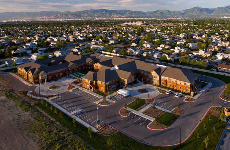 Aerial view of a large senior living facility building with multiple wings, surrounded by parking lots and landscaped areas, set within a suburban neighborhood with mountains in the background.