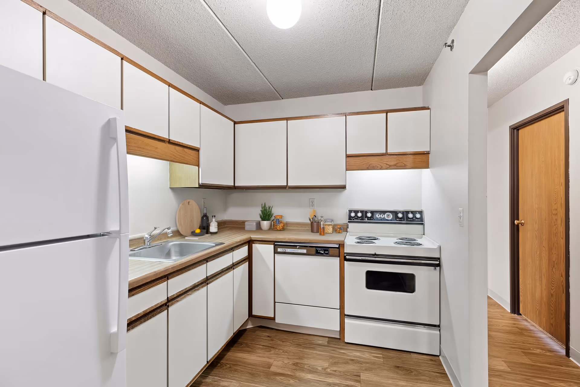 A compact kitchen with white cabinets and wood trim, a white refrigerator, a stainless steel sink, a white dishwasher, and a white electric stove. The kitchen has wood flooring and a ceiling light. There are small decorative items on the countertop including a small plant and jars.