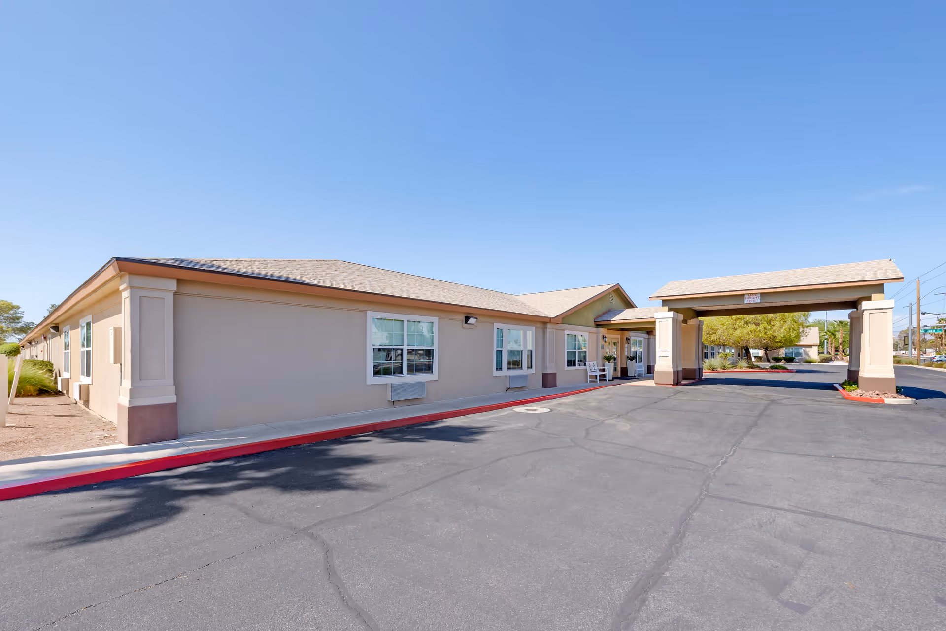 Exterior view of a single-story building with beige walls and a brown roof under a clear blue sky. The building has multiple windows and a covered entrance supported by columns. There is a paved driveway with red curbs and some landscaping with trees and plants near the entrance.