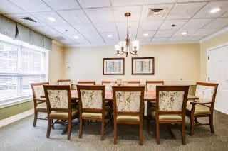 A well-lit dining room with a long wooden table surrounded by ten upholstered chairs with floral patterns. The room has beige walls, a chandelier hanging above the table, two framed pictures on the wall, and a large window with blinds and a valance allowing natural light to enter.