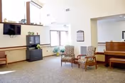 A bright common area in a nursing home with two patterned armchairs, a piano, a black TV mounted on the wall, a black cabinet, and large windows letting in natural light. The room has beige walls and carpeted flooring.