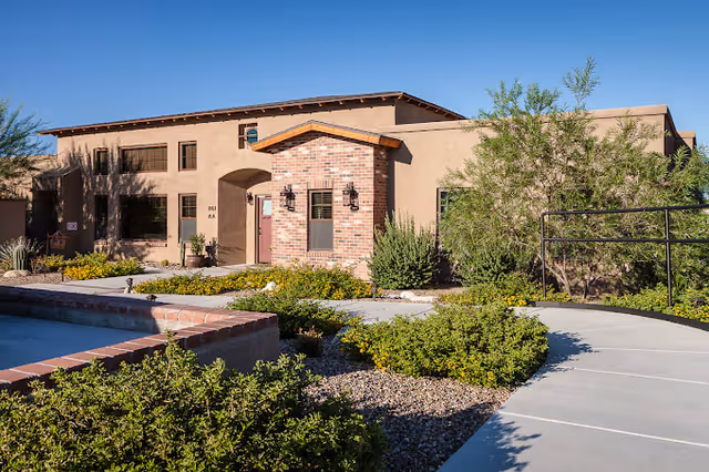 Exterior view of a single-story senior living facility building with a beige stucco finish and a small brick entrance. The building is surrounded by desert landscaping with bushes, gravel, and a paved walkway leading to the entrance. The sky is clear and blue.