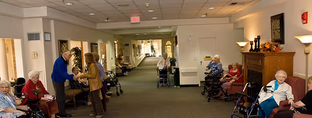 A hallway in a retirement community with elderly residents sitting on chairs along the walls, some using walkers. Two people are standing and conversing near the left side. The hallway is well-lit with ceiling lights and has a carpeted floor. There is a fireplace with decorative items on the right side.
