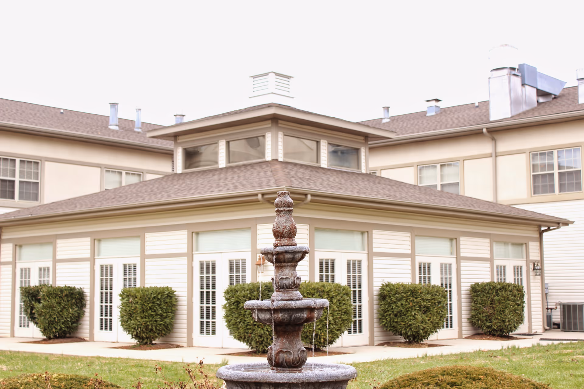Exterior front of a senior living building with a multi-tiered fountain in the foreground and trimmed shrubs along the windows.