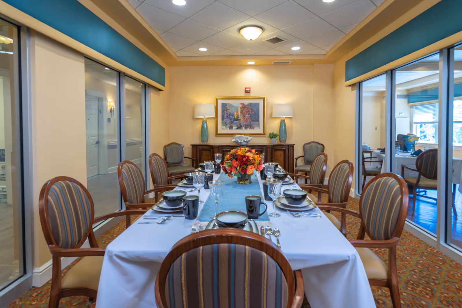 Formal dining room with a long table set for a meal, a floral centerpiece, and striped wooden chairs in a senior living facility.