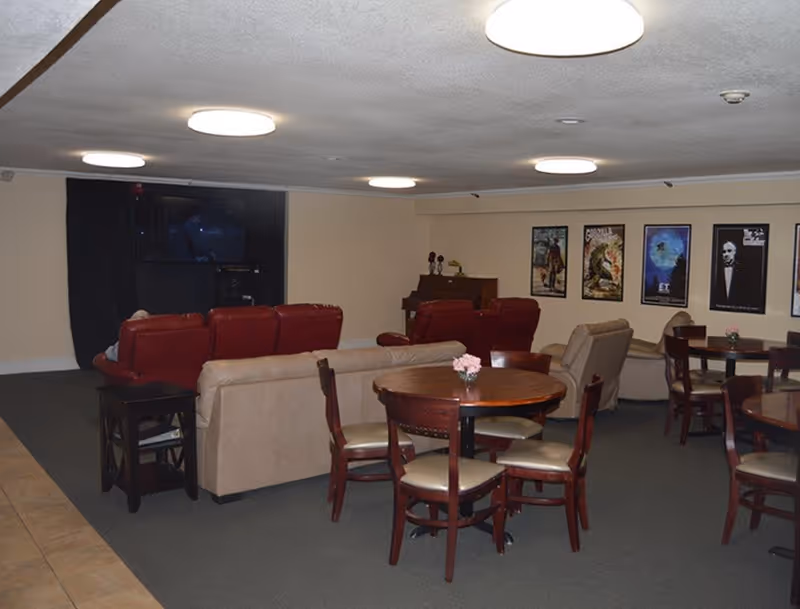 A common area with multiple round wooden tables and chairs, beige and red sofas facing a television mounted on the wall, movie posters on the wall, and a piano in the corner.