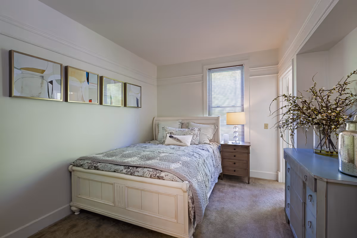 A bright bedroom with a bed, patterned bedding, a nightstand with a lamp by a window, dresser and wall art above the bed.