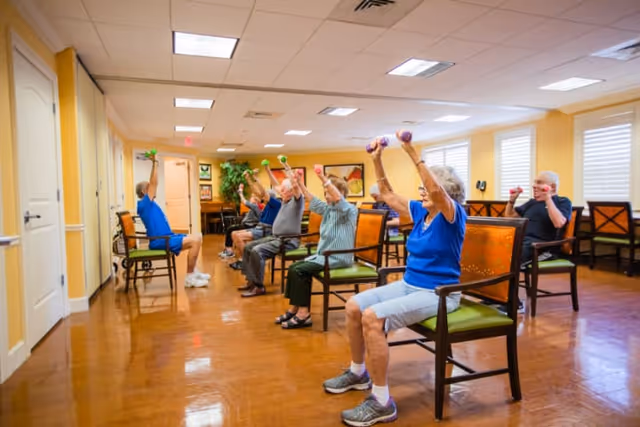A group of elderly individuals seated in a well-lit room participating in a seated exercise class, each holding small dumbbells and raising their arms above their heads.