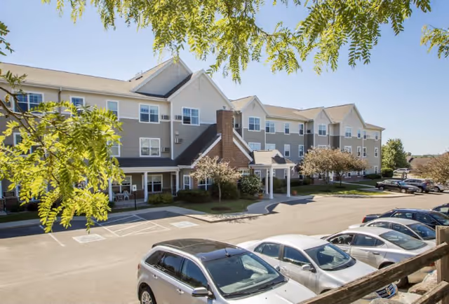 Exterior view of a multi-story senior living facility building with beige and gray siding, multiple windows, and a covered entrance. Several cars are parked in the parking lot in front of the building, and green trees with leaves frame the top of the image under a clear blue sky.