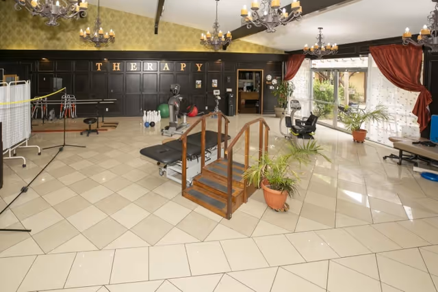 A spacious therapy room with tiled floors and high ceilings featuring chandeliers. The room contains various physical therapy equipment including a therapy table with wooden handrails and steps, exercise balls, a stationary bike, and a set of bowling pins. There are large windows with red curtains allowing natural light to enter, and several potted plants are placed around the room. The word 'THERAPY' is displayed on a dark wood-paneled wall in the background.