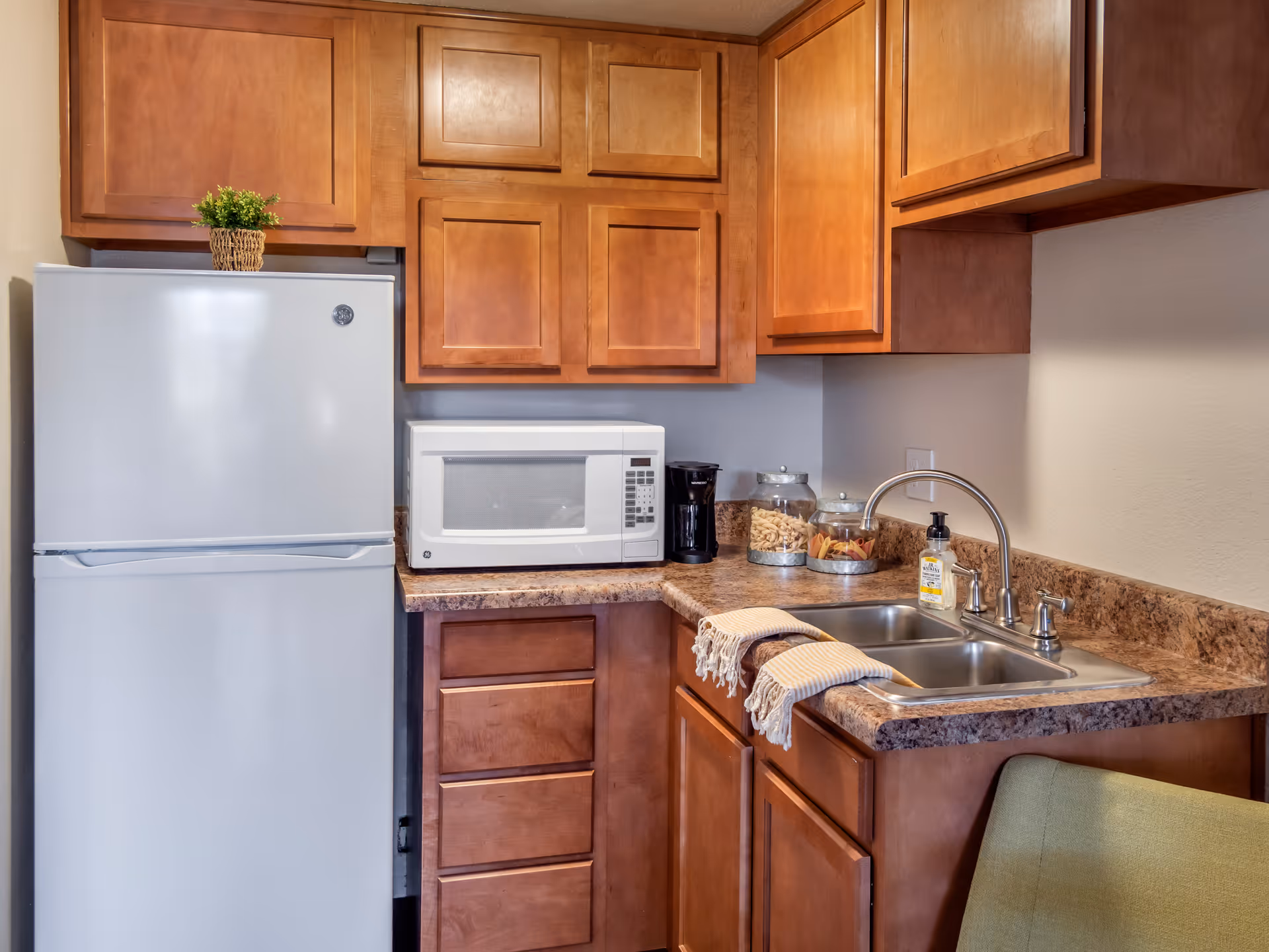 A small kitchen area with wooden cabinets, a white refrigerator, a white microwave, a coffee maker, two glass jars with pasta, a double sink with a faucet, and two striped dish towels hanging over the sink edge. There is a green chair partially visible in the foreground.