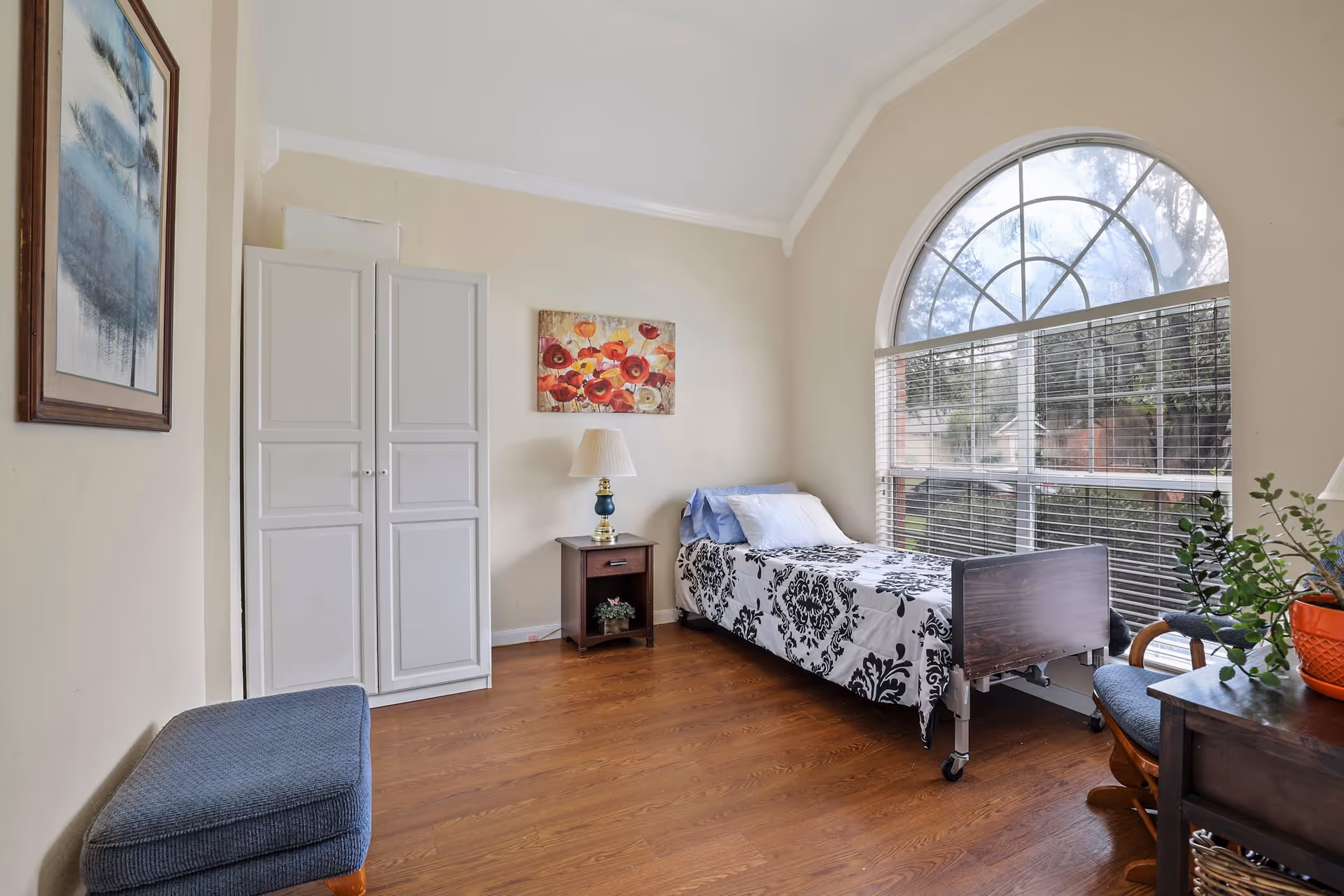 A bright and cozy bedroom with a single bed featuring a black and white patterned bedspread. The room has wooden flooring, a large arched window with blinds letting in natural light, a small wooden nightstand with a lamp, a white wardrobe, a blue cushioned bench, a wooden chair, and framed artwork on the walls.