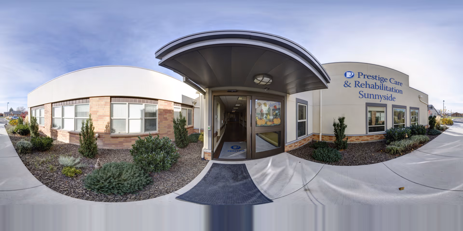 Exterior view of the entrance to Prestige Care & Rehabilitation Sunnyside, showing a curved canopy over the doorway, windows along the building, and landscaped bushes and plants around the walkway.