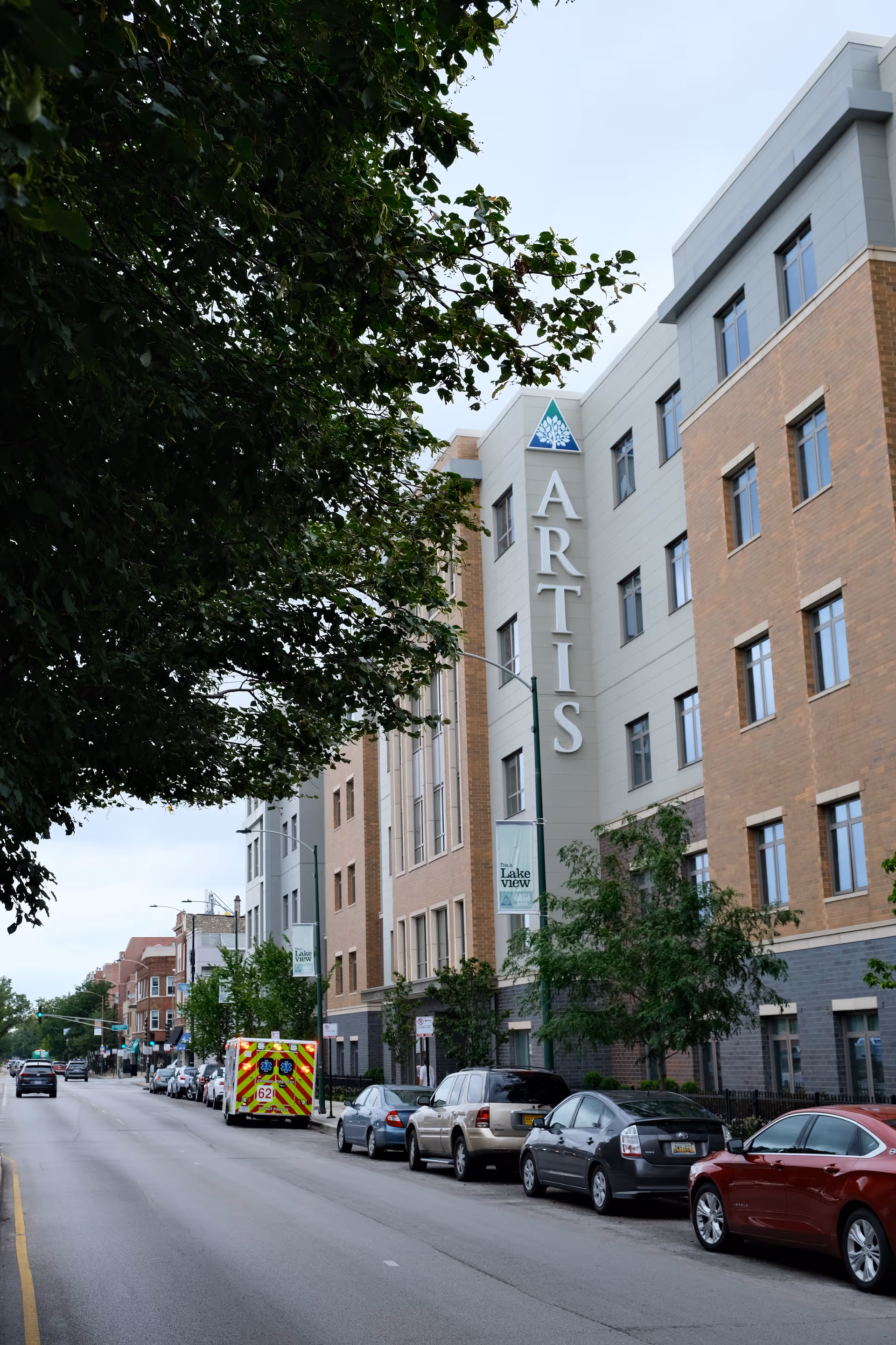 Street view of the Artis Senior Living building showing a tall façade with vertical 'ARTIS' signage, trees, and parked cars along the street.
