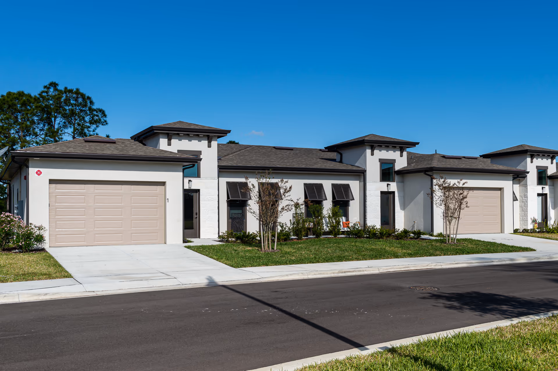 Exterior view of a modern single-story residential building with beige garage doors, white walls, and dark shingled roofs under a clear blue sky. The building is surrounded by a well-maintained lawn and small trees, with a paved road in the foreground.