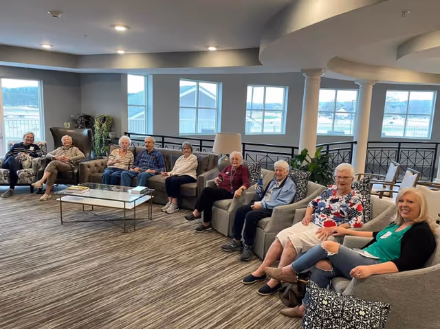 A group of elderly people sitting comfortably in a spacious, well-lit common area with large windows, modern furniture including armchairs and sofas, a glass coffee table, and indoor plants. The setting appears warm and inviting, with a carpeted floor and ceiling lights.