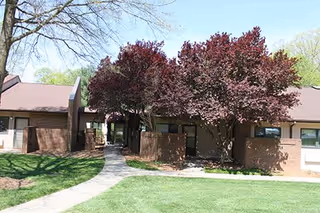 Exterior view of a single-story brick building with a pathway leading through a courtyard area. There are trees with reddish leaves and green grass surrounding the building under a clear blue sky.