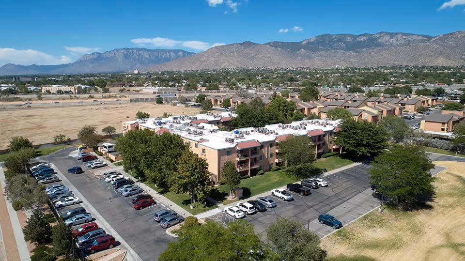 Aerial view of Sandia Vista Senior Living facility showing multiple apartment buildings with white roofs, surrounded by trees and parking lots filled with cars. In the background, there is a wide expanse of land and mountain range under a clear blue sky.