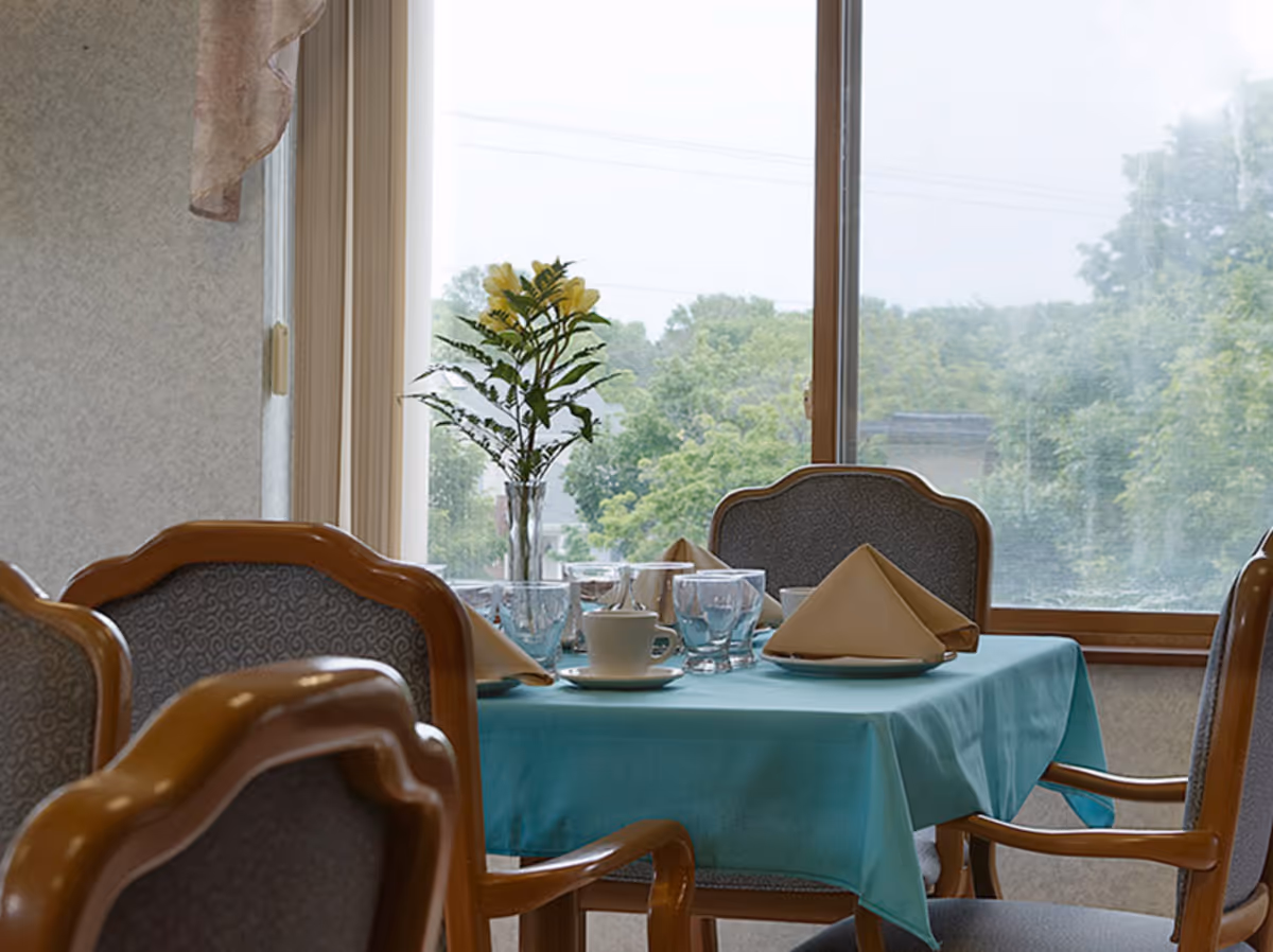 A dining table set with a turquoise tablecloth, beige folded napkins, cups, glasses, and a vase with yellow flowers, surrounded by wooden chairs with cushioned seats and backs, next to a large window showing green trees outside.
