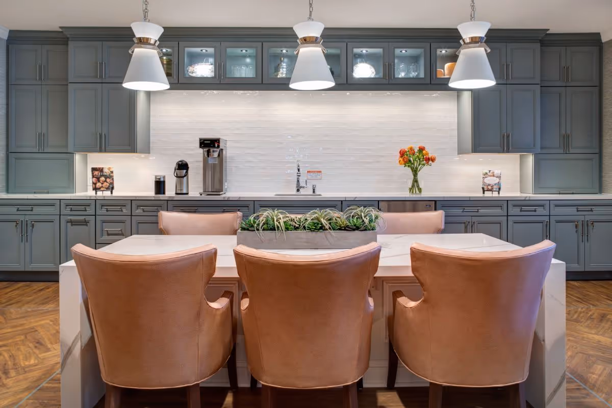Modern communal kitchen with a marble island, three leather chairs, pendant lights, gray cabinetry and a coffee station.