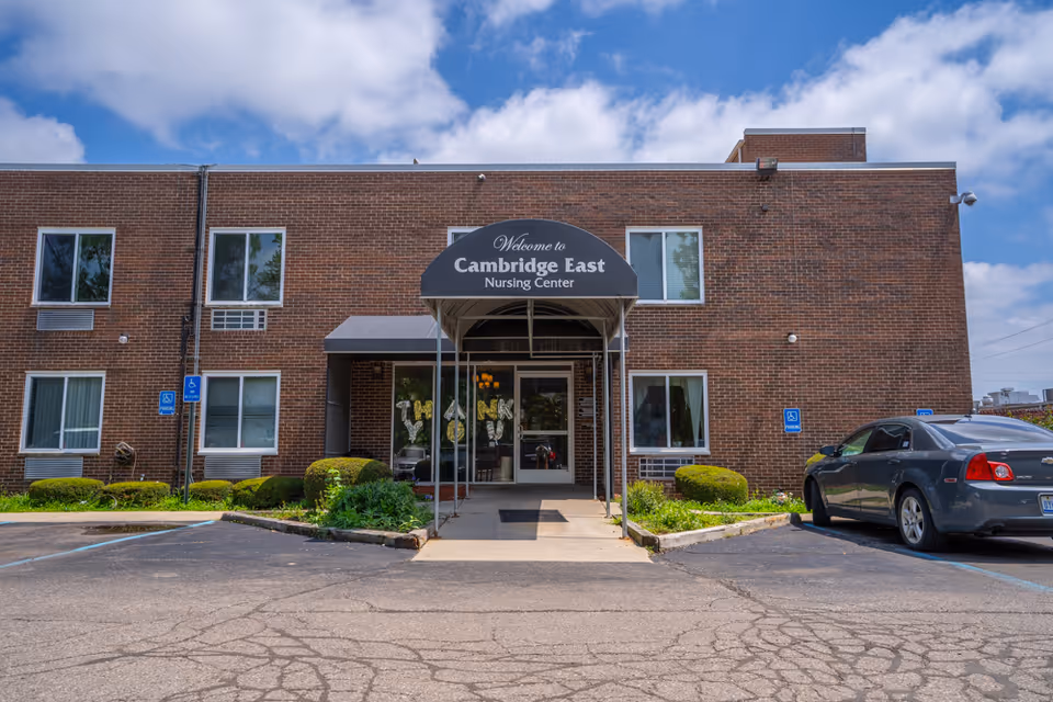 Front exterior view of a two-story brick nursing center building with a covered entrance canopy that reads 'Welcome to Cambridge East Nursing Center'. There are several windows, some bushes and plants along the front, a parking lot with a few cars, and a clear blue sky with some clouds.