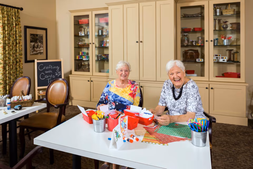 Two elderly women sitting at a table in a crafting room, smiling and working on card-making projects with various craft supplies like scissors, glue sticks, and colored paper. Behind them are beige cabinets with glass doors displaying more craft materials. A chalkboard sign reads 'Crafting with Cards Today.'