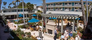 Outdoor patio area at The Village at The Palms senior living facility with tables, chairs, umbrellas, and a pergola with a fireplace. Several people are seated and standing, enjoying the sunny weather surrounded by palm trees and a multi-story building in the background.