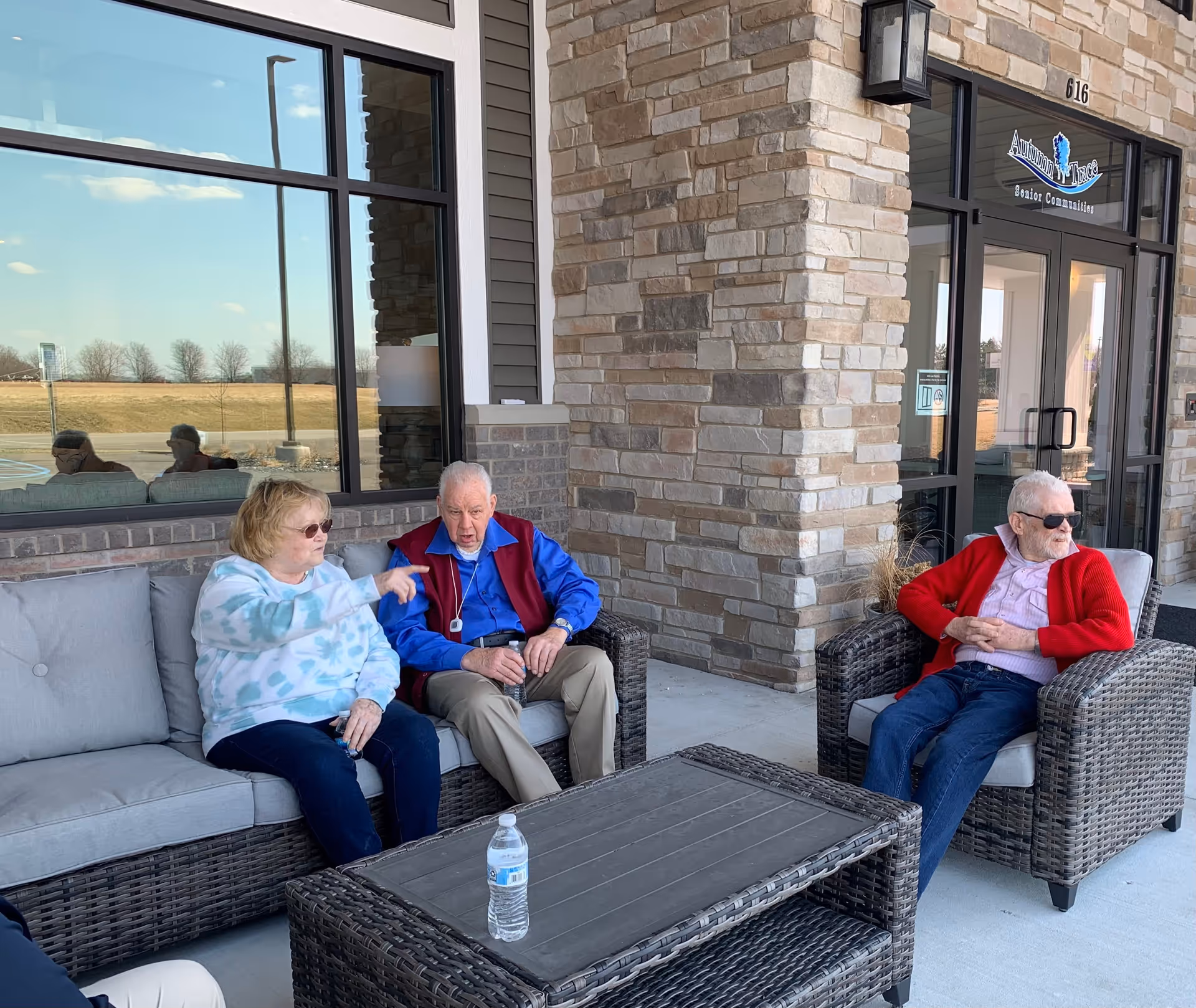 Three elderly people sitting on outdoor wicker furniture with cushions in front of a stone building. Two are seated on a sofa, one wearing a light blue patterned sweatshirt and the other in a blue shirt with a red vest. The third person is sitting on a single chair wearing a red cardigan and sunglasses. A water bottle is on the table in front of them. The building has large windows and a glass door with a sign above reading 'Autumn Trace Senior Communities'.
